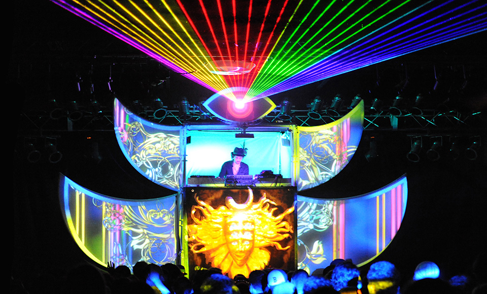 Historical Shpongletron stage photograph: a performer between curved projection surfaces, a glowing mask below and rainbow lasers from an eye above